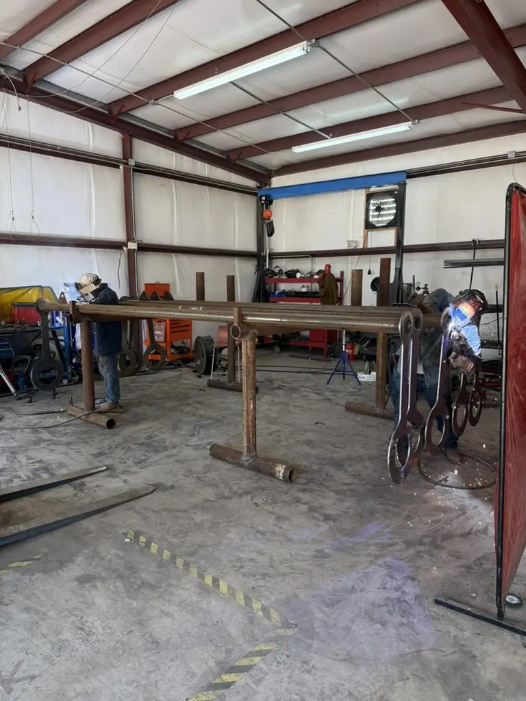 Long steel frame on stands across the shop floor with a welder at the far end